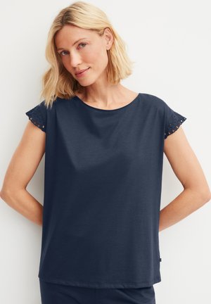 Blonde vrouw met schouderlang haar, gekleed in een marineblauw kort mouwtopje met ajourkant aan de mouwen, poserend tegen een witte achtergrond.