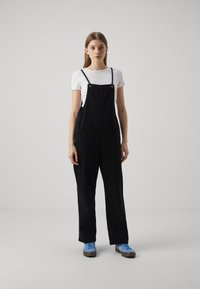 Sorte bomullsdresser med justerbare stropper, to frontlommer og avslappet passform, kombinert med en hvit t-skjorte og blå sneakers.