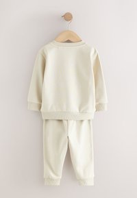 Kreemivärvi pusad ja sobivad püksid pehmest kangast, millel on soonikkoes mansetid ja lõdvestunud lõige, riputatud puidust riidepuule.