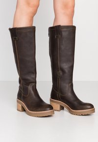 Tmavě hnědé kožené kozačky nad kolena s texturovaným povrchem, opatřené bočním zipem a dřevěným blokovým podpatkem s gumovou podrážkou.