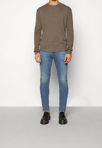 Hombre vistiendo una camisa de manga larga verde oliva, jeans ajustados azul claro y zapatos negros con cordones, de pie contra un fondo blanco y gris liso.