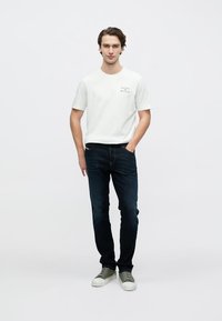 Bela bombažna majica s majhnim natisnjenim logotipom na prsih, kombinirana s temno modrimi kavbojkami in sivimi supergami. Preprosta in priložnostna obleka.