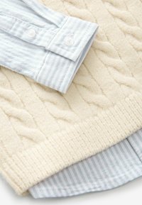 Un pull en maille torsadée couleur crème superposé sur une chemise rayée bleu clair et blanche, avec des poignets boutonnés et des textures douces.