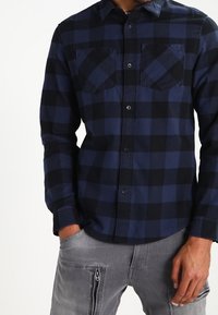 Chemise en flanelle à carreaux bleus et noirs avec col boutonné, deux poches poitrine et manches longues, portée avec un pantalon cargo gris.