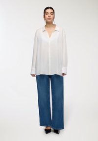 Camicia bianca a maniche lunghe con colletto, abbinata a jeans in denim blu scuro a gamba larga. Tessuto leggero e liscio.