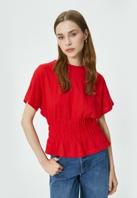 Top rosso corto con maniche ampie e vita arricciata, abbinato a jeans blu. Tessuto liscio e leggero con una vestibilità rilassata.
