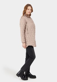 Beige gewatteerde jas met een gestructureerde kraag, gecombineerd met zwarte jeans en stevige zwarte laarzen. De jas heeft een relaxte pasvorm en elastische manchetten.