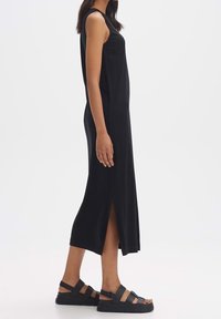 Robe noire sans manches avec une fente latérale, en tissu doux. Associée à des sandales noires à brides avec une semelle épaisse.