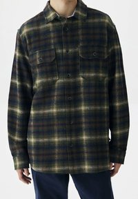 Camisa de franela con un patrón de cuadros en verde, azul marino y marrón. Dos bolsillos en el pecho, cierre de botones y mangas largas. Textura suave.