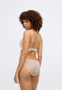 Γυμνός ασύρματος σουτιέν με ρυθμιζόμενες τιράντες και διάφανο πίσω μέρος, συνδυασμένο με αντίστοιχα διάφανα εσώρουχα πλήρους κάλυψης. Λείος υφή και ελάχιστος σχεδιασμός.