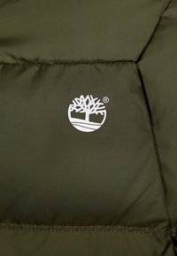 Giacca puffer verde oliva in tessuto con texture trapuntata; presenta un logo bianco di un albero all'interno di un contorno circolare sulla zona del torace.