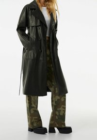 Žena v dlouhém černém koženém trench coatu přes bílý crop top, maskáčových kalhotách a černých platformových botách, stojící se založenýma rukama v kapsách.