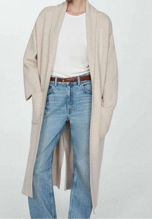 Persoon die een lange beige open cardigan draagt, een wit T-shirt, een bruine riem en lichtblauwe hoog getailleerde spijkerbroek tegen een effen achtergrond.