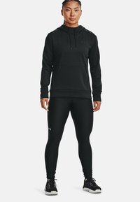 Felpa nera con cappuccio, dotata di tasca frontale e logo sul petto, abbinata a leggings neri. Materiale liscio, tessuto attillato. Sneakers nere.