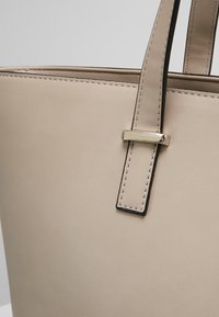 Beige læder tote taske med en glat tekstur, der har en rektangulær form, elegant sort syning og en metallisk spænde detalje på remmen.