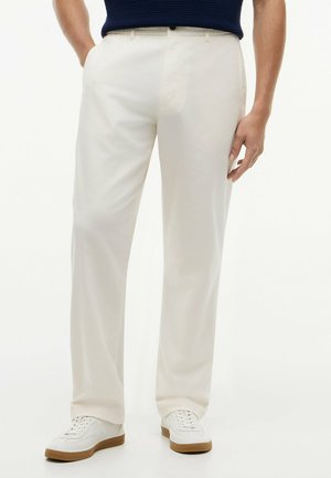 Mann trägt weiße gerade geschnittene Hose, navyblaues geripptes Shirt und weiße Sneaker mit braunen Sohlen, steht mit einer Hand in der Tasche.