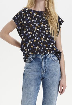 Schwarze Bluse mit kurzen Ärmeln, die ein floralem Muster in Blau, Weiß und Gelb aufweist. Kombiniert mit hochgeschnittenen hellblauen Jeans.
