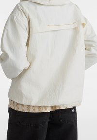 Vit vindjacka med en strukturerad yta, med en bakficka med kontrasterande dragkedja. Kombinerad med en mönstrad beige skjorta och svarta byxor.