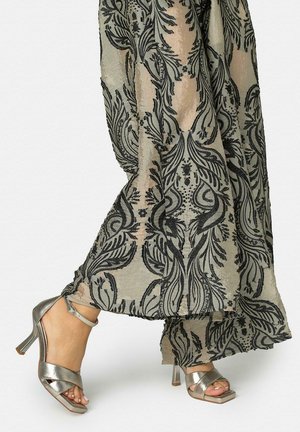 Mujer con pantalones anchos transparentes con estampado floral y sandalias de tacón alto plateadas metálicas con tiras en el tobillo caminando sobre un suelo blanco.