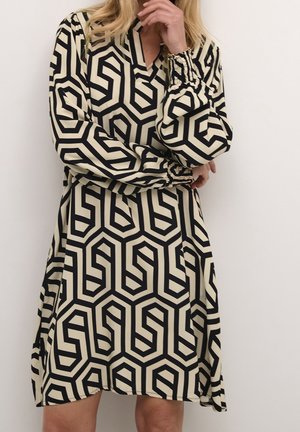 Nainen pukeutuneena musta-beigeen geometrisesti kuvioituun mekkoon, jossa on pitkät hihat ja joustavat kalvosimet, poseeraa siten, että yksi käsi on leukansa lähellä.