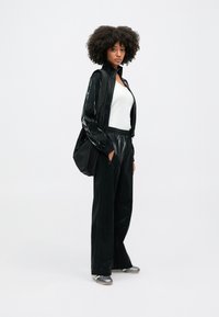 Μαύρο, γυαλιστερό φόρμα με ζακέτα με φερμουάρ και παντελόνι με ελαστική μέση. Το μοντέλο φοράει λευκό μπλουζάκι και μεταλλικά αθλητικά παπούτσια, σε φόντο λευκό.