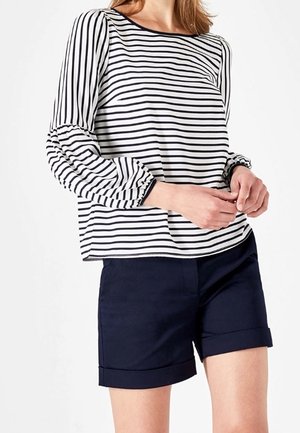 Vrouw die een wit shirt met lange mouwen en zwarte horizontale strepen draagt, en marineblauwe korte broek met omslag, handen losjes gekruist op heuphoogte.