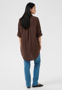 Barna hosszú ujjú ing felhajtott mandzsettával, egyenes szegéllyel és puha textúrával, kék farmerrel és fekete lapos cipővel párosítva.