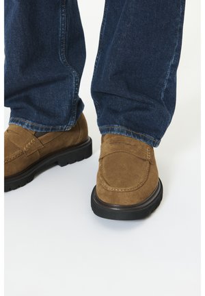 Brune ruskind loafers med sorte såler båret med mørkeblå denimjeans stående på en hvid overflade.