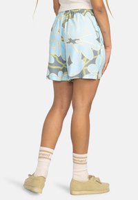 Shorts bleus à motif floral avec une taille élastique, dotés de fentes latérales. Portés avec des chaussures beiges et des chaussettes blanches.