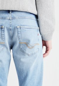 Jeans en denim bleu clair avec une coupe décontractée, dotés de deux poches arrière avec des coutures jaunes et d'un logo brodé sur une poche.