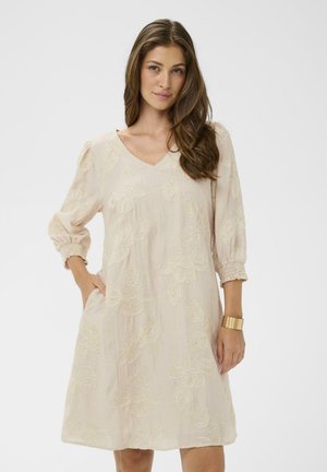 Femme aux longs cheveux bruns portant une robe florale beige clair à manches trois-quarts et un bracelet en or, posant avec une main dans la poche.