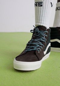 Brown suede high-top sneakers with white sole, white side stripe, and blue speckled laces. Worn with white ribbed socks.