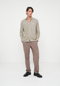 Lichtbeige shirt met zwart cirkelpatroon, lange mouwen en kraag, gecombineerd met lichtbruine broek en zwarte schoenen.
