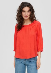 Blouse en tissu rouge froissé avec des manches trois-quarts, un col volanté et une coupe ample, associée à un jean bleu clair.
