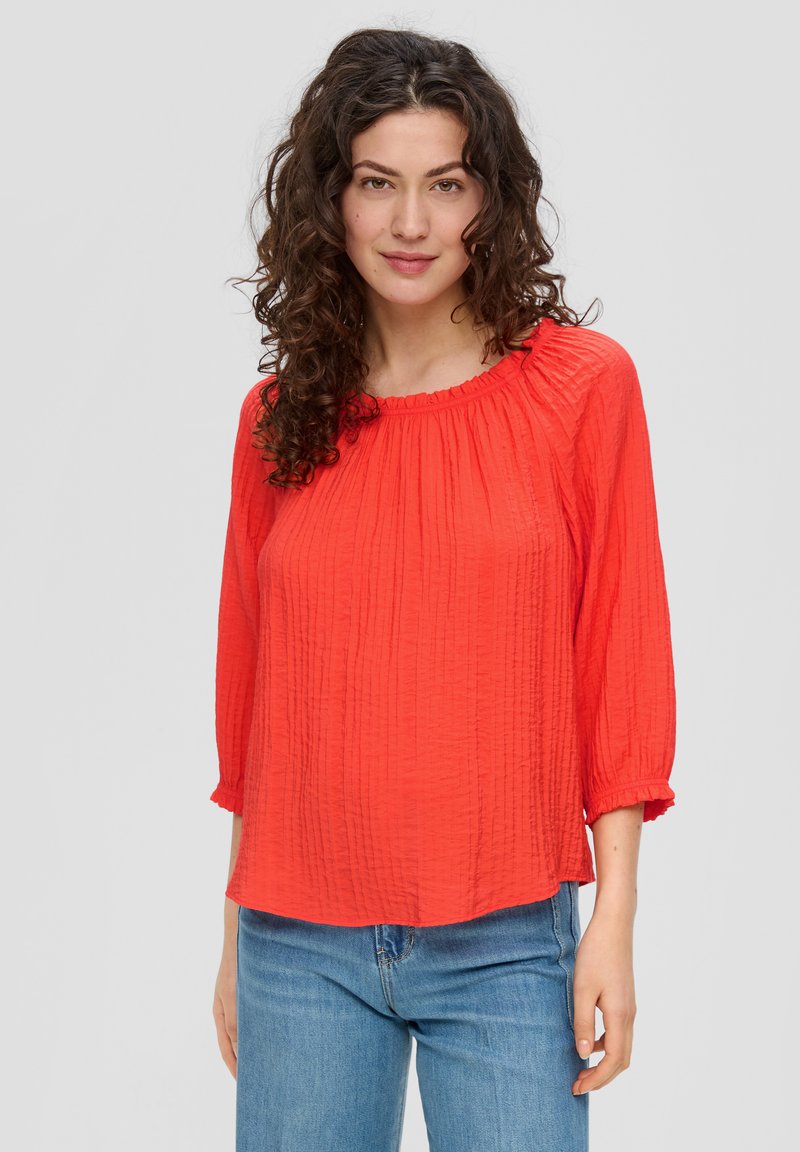 Blouse en tissu rouge froissé avec des manches trois-quarts, un col volanté et une coupe ample, associée à un jean bleu clair.