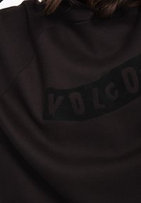 Mustapaita, jossa on sileä pinta, ja takana on iso tummalla kirjoitettu logo, joka lukee "VOLCOM" paksuilla kirjaimilla.
