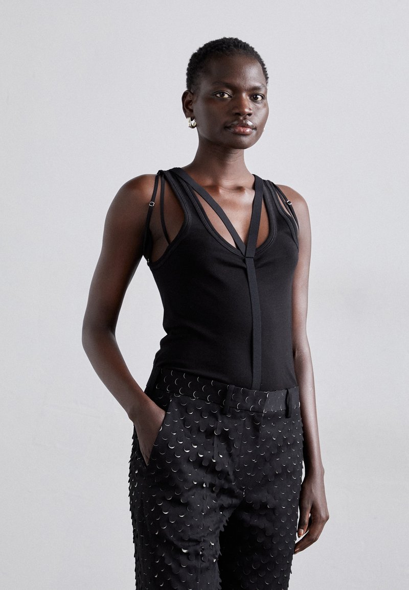 Fekete ujjatlan felső mély V-kivágással és bonyolult pántokkal, párosítva textúrált fekete nadrággal, amely félkör alakú mintázattal rendelkezik.