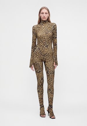 Vrouw staand gekleed in een volledige luipaardprint bodysuit met lange mouwen en hoge halslijn, gecombineerd met zwarte, bandjes high-heeled sandalen.