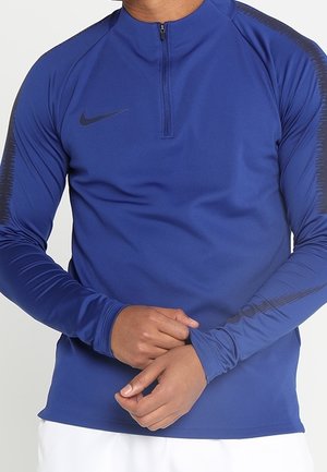 Man wearing a blue long-sleeve Nike sports top with a quarter zip, adjusting the cuff, white pants visible at waist level.
