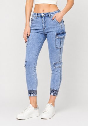 Kvinde iført lyseblå denim cargo joggerbukser med elastiske manchetter og hvide sneakers, stående mod en ensartet baggrund.