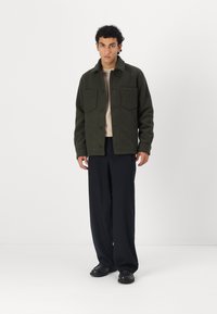 Donkergroene wollen blend jas met twee borstzakken, knoopsluiting en een overhemdkraag, gedragen over een lichtbeige trui en zwarte broek.