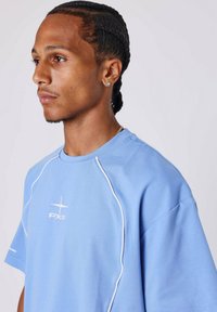 Joven con cabello trenzado y un pendiente de diamante, vistiendo una camiseta PXP de color azul claro con ribete blanco, mirando hacia la izquierda sobre un fondo liso.