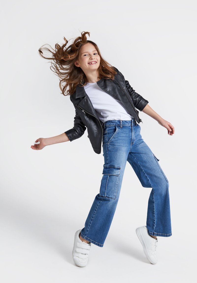 Czarna skórzana kurtka, biała koszulka, niebieskie szerokie spodnie cargo z kieszeniami i białe trampki; model w dynamicznej pozie na jednolitym tle.