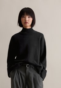 Μαύρο πουλόβερ με καλλιεργημένο λαιμό και μανσέτες, φορεμένο με γκρι παντελόνι ψηλόμεσο και μαύρη ζώνη. Μαλακή υφή.