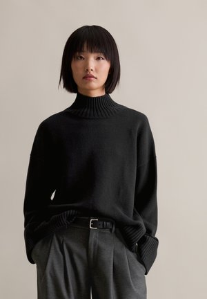 Schwarzer Rollkragenpullover mit Rippkragen und -manschetten, getragen mit grauen hochgeschnittenen Hosen und einem schwarzen Gürtel. Weiche Textur.