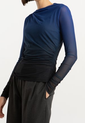 Kobieta ubrana w dopasowany, marszczony, granatowo-czarny ombre top z długim rękawem i przezroczystymi rękawami oraz luźne ciemnoszare spodnie, ręka w kieszeni.