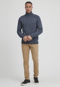 Mornarsko pleteno pulover z visokim ovratnikom, teksturiran z lahkimi pikami, v kombinaciji s svetlo bež slim-fit hlačami in črnimi supergami.