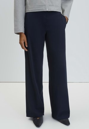 Person trägt lockere dunkelblaue Hose und schwarze spitze Schuhe, mit einer Hand in der Tasche und grauer Jacke, die an der Taille sichtbar ist.
