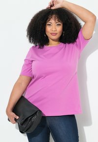 T-shirt a maniche corte, con collo tondo, in tessuto rosa brillante, abbinato a una borsa a tracolla nera. Il modello indossa jeans scuri.