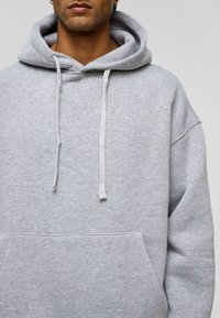 Lichtgrijze hoodie met een grote voorzak, koorden en een ontspannen pasvorm. De stof lijkt zacht en gestructureerd.